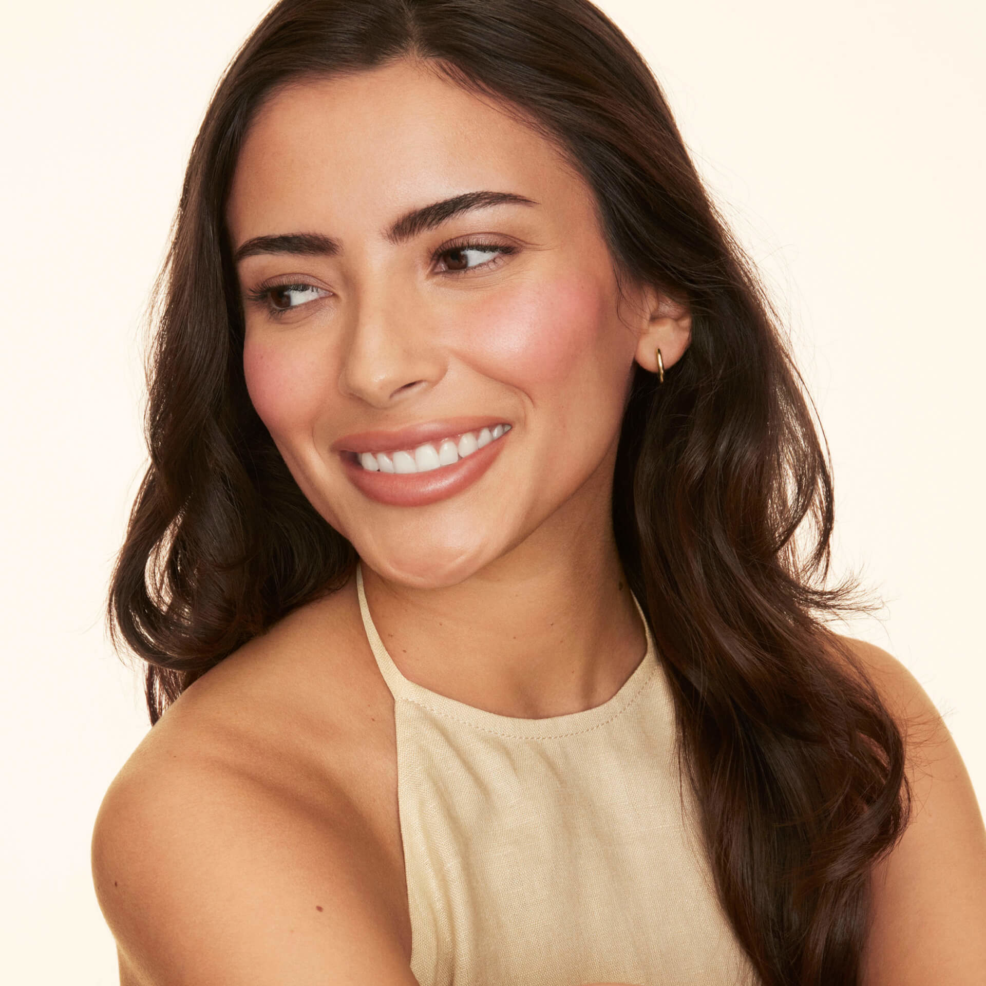 Woman with long brown hair and natural makeup, wearing a sleeveless beige top, smiling and looking slightly to the side against a light background.